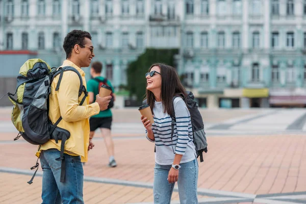 Glückliche interrassische Freunde Mann und Frau, die einander anschauen und mit Rucksäcken und Coffee-to-go lächeln — Stock Photo