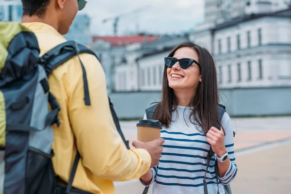 Mujer sonriente con gafas de sol mirando a un amigo bi-racial con taza de papel de café y mochila - foto de stock