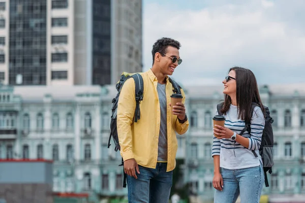 Mulher olhando para amigo bi-racial e sorrindo com xícara de papel de café e mochila — Fotografia de Stock