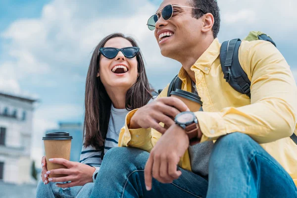 Amigos interraciales con mochilas sentadas en la calle y tomando café - foto de stock