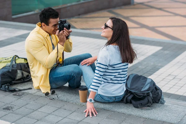 Hombre bi-racial sentado en las escaleras y tomando fotos de mujer en gafas de sol con mochila - foto de stock