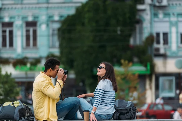Двурасовый мужчина сидит и фотографирует женщину в солнечных очках с рюкзаком — стоковое фото