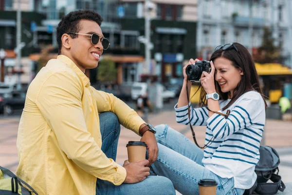 Woman sitting and taking photo of multiracial man in sunglasses and coffee cup — Stock Photo