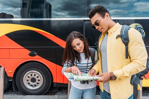 Woman with digital camera pointing with finger on map with mixed race man near travel bus — Stock Photo