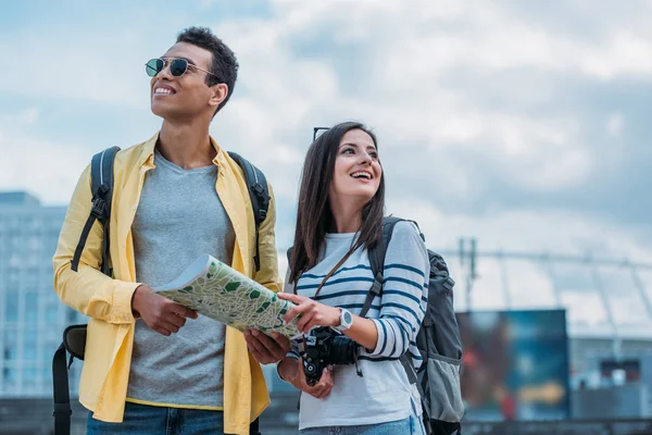 Mujer con cámara digital y amigo de raza mixta sosteniendo mapa y sonriendo - foto de stock