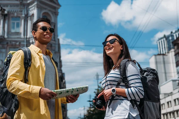 Femme en lunettes de soleil avec appareil photo numérique proche ami bi-racial avec carte et sac à dos — Photo de stock