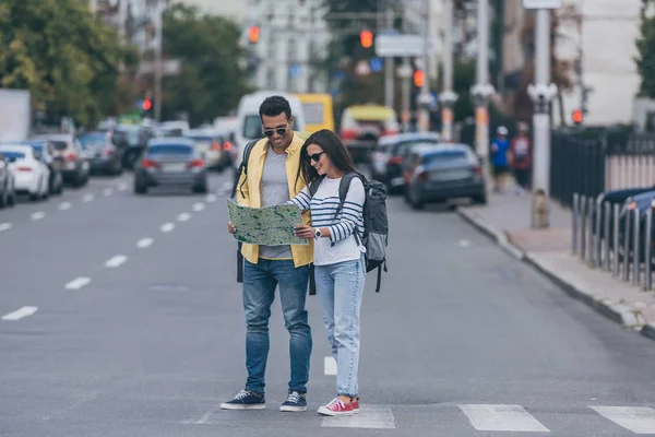 Woman and multiracial friend with backpacks holding city map on crosswalk — Stock Photo