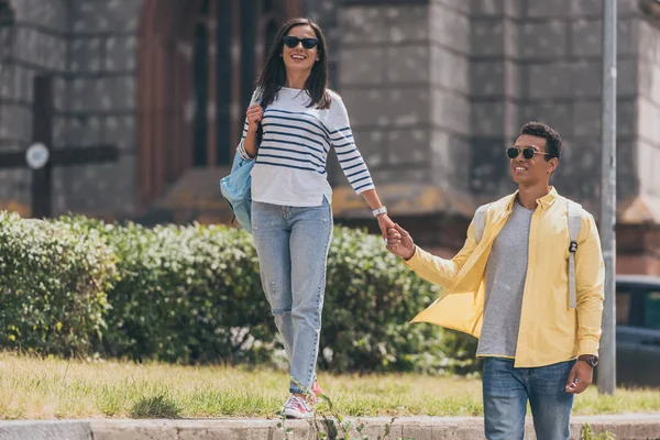 Hombre multiracial en gafas de sol con mujer cogida de la mano y caminando - foto de stock