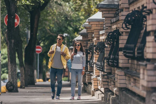 Homme bi-racial en lunettes de soleil près d'une femme marchant avec un sac à dos — Photo de stock