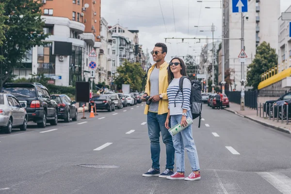 Mujer sosteniendo mapa y de pie en la carretera cerca de bi-racial amigo con mochila - foto de stock