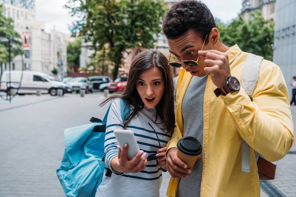 Hombre bi-racial en gafas de sol y mujer mirando en el teléfono inteligente y sorprendente - foto de stock