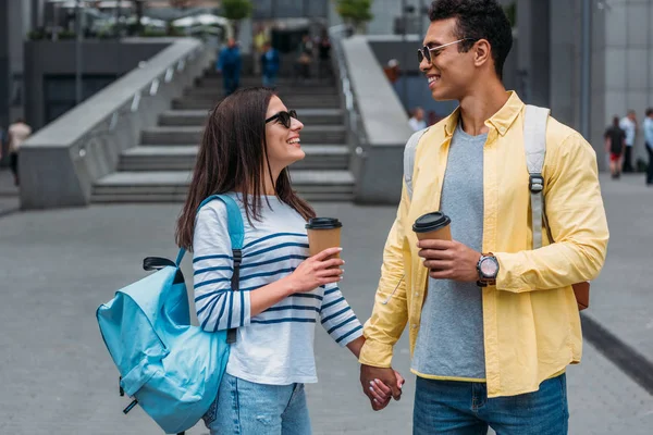 Uomo bi-razziale in occhiali da sole e donna con tazza di carta che si tiene per mano e si guarda l'un l'altro — Foto stock
