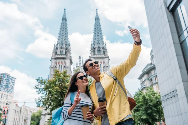 KYIV, UCRANIA - 8 de julio de 2019: Mujer cerca de hombre bi-racial en gafas de sol tomando selfie en smartphone - foto de stock