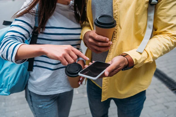 Cropped view of bi-racial man with paper cup holding smartphone and woman pointing with finger — Stock Photo