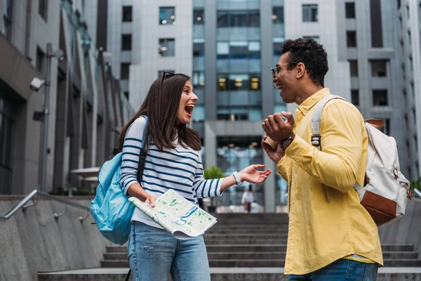 Smiling bi-racial man with backpack and woman holding map near stairs — Stock Photo
