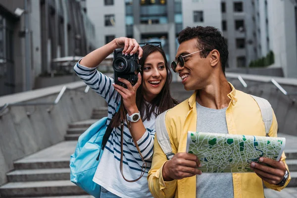 Mujer tomando fotos en cámara digital y mirando bi-racial amigo sosteniendo mapa - foto de stock