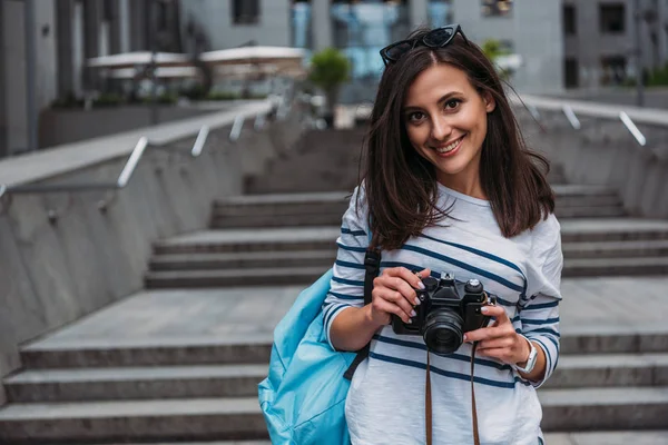 Mujer en casual con mochila sosteniendo cámara digital y sonriendo - foto de stock
