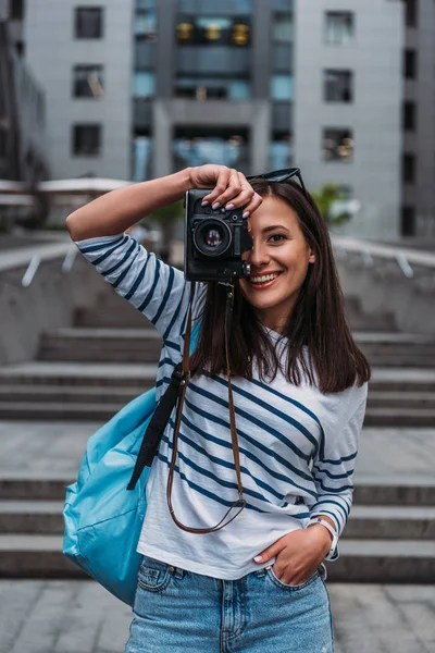 Fille heureuse prenant des photos sur appareil photo numérique et debout avec la main dans la poche à l'extérieur — Photo de stock