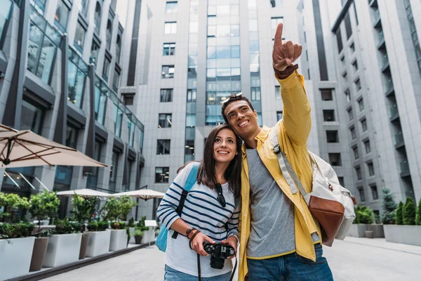 Счастливая девушка с цифровой камерой рядом веселый смешанной расы мужчина указывая пальцем — стоковое фото