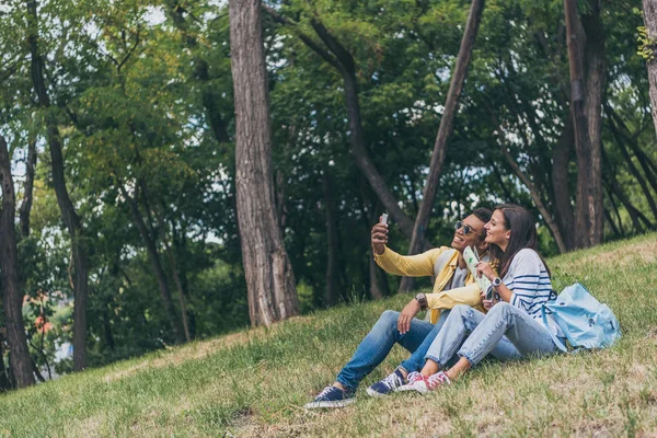 Щасливий змішана раса чоловік бере селфі з веселою дівчиною, сидячи на траві — стокове фото