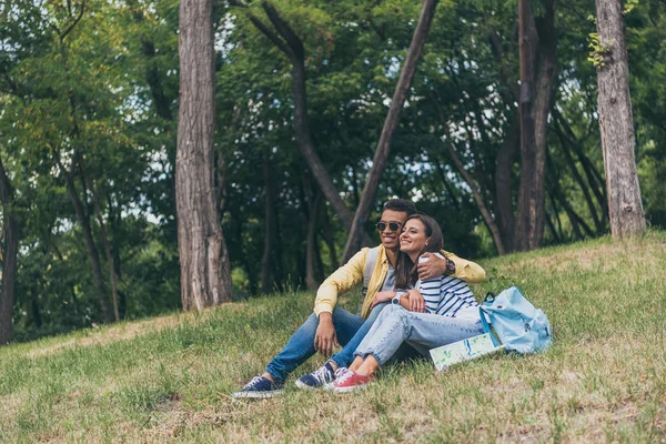 Glücklich gemischte Rasse Mann umarmt fröhliche Mädchen, während sie auf Gras sitzen — Stockfoto