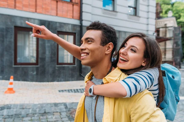 Glückliches Mädchen umarmt fröhlich gemischten Rassenmann und zeigt mit dem Finger in der Nähe Gebäude — Stockfoto