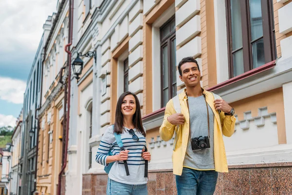 Happy girl touching backpack while walking with mixed race man near buildings in city — Stock Photo