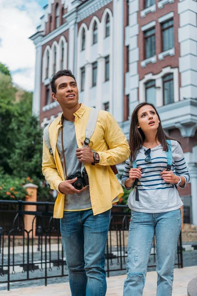 Surprised girl touching backpack while standing with mixed race man near building — Stock Photo