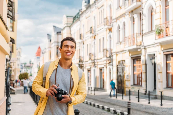 Allegro uomo di razza mista sorridente mentre tiene la fotocamera digitale vicino agli edifici — Foto stock