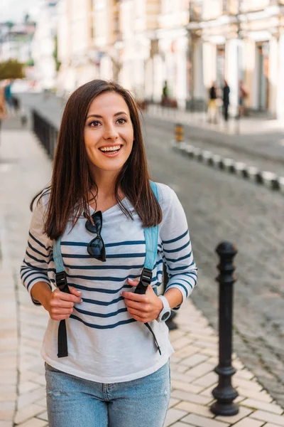 Happy girl touching backpack and smiling near near building — Stock Photo