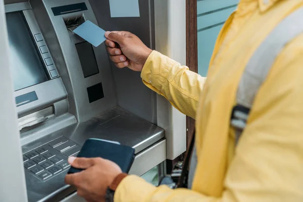 Vista recortada de hombre de raza mixta poner la tarjeta de crédito en el cajero automático mientras sostiene la cartera - foto de stock