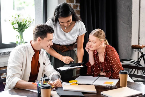 Three multiethnic friends sitting at table and looking at calculator in apartment — Stock Photo