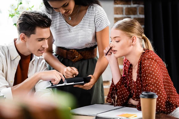 Tres amigos multiétnicos sentados en la mesa y mirando la calculadora en el apartamento - foto de stock