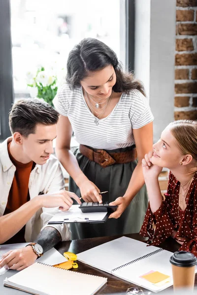 Three multiethnic friends sitting at table and looking at calculator in apartment — Stock Photo