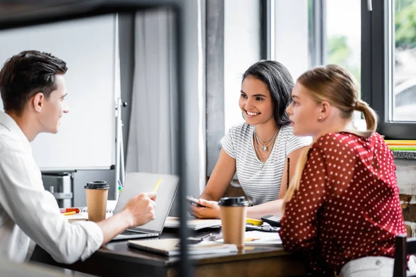 Tres amigos multiétnicos sentados en la mesa y hablando en el apartamento - foto de stock