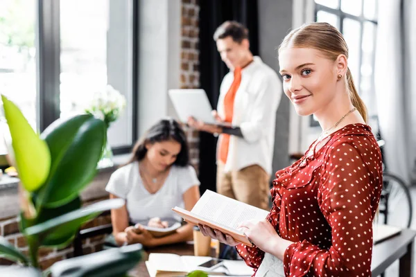 Selective focus of attractive student holding book and looking at camera — Stock Photo