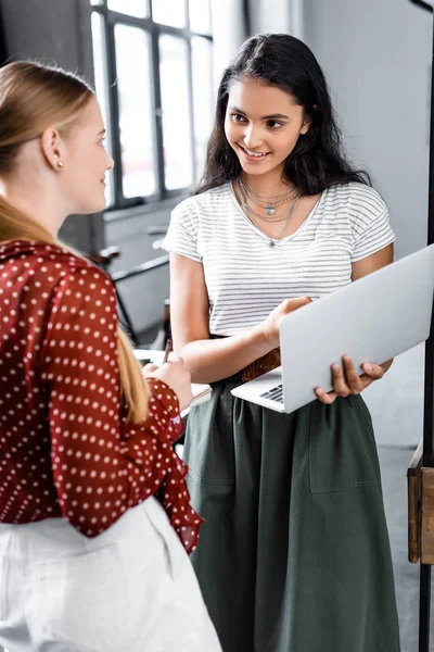 Atractivos estudiantes multiétnicos sonriendo y sosteniendo portátil en apartamento — Stock Photo