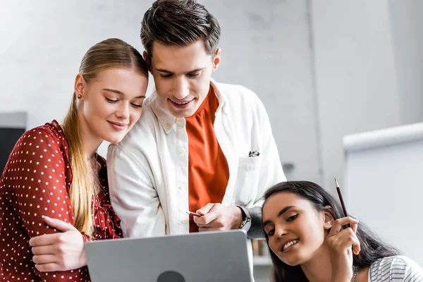 Multiethnic students smiling and looking at laptop in apartment — Stock Photo