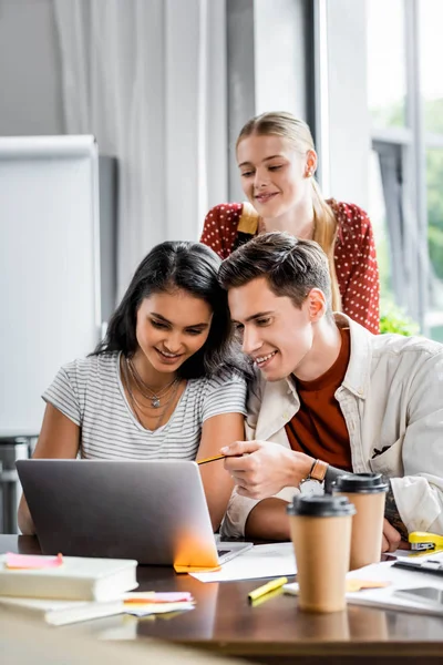 Estudantes multiétnicos sorrindo e olhando para laptop no apartamento — Fotografia de Stock