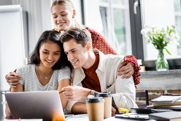 Estudantes multiétnicos sorrindo e olhando para laptop no apartamento — Fotografia de Stock