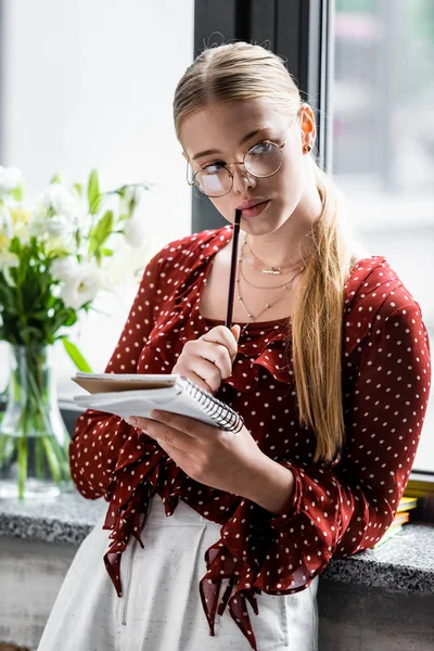 Attractive and pensive student in glasses holding notebook and pencil in apartment — Stock Photo