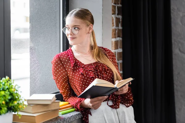 Attraente e sorridente studente in bicchieri tenendo libro in appartamento — Foto stock