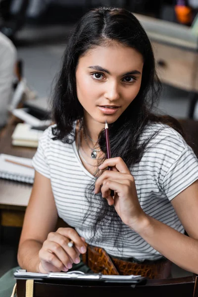 Attractive and bi-racial student holding pencil and looking at camera — Stock Photo