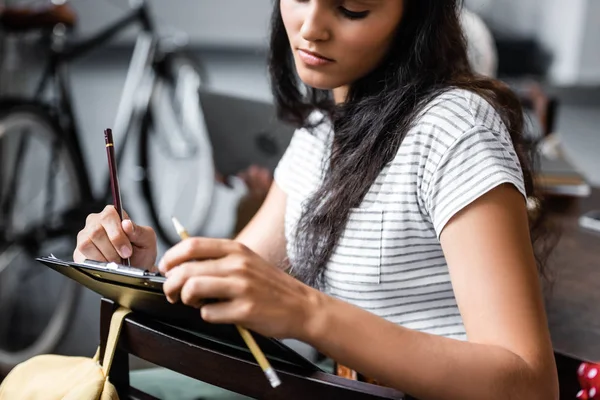 Привлекательный и двурасовый студент, пишущий карандашом в квартире — стоковое фото