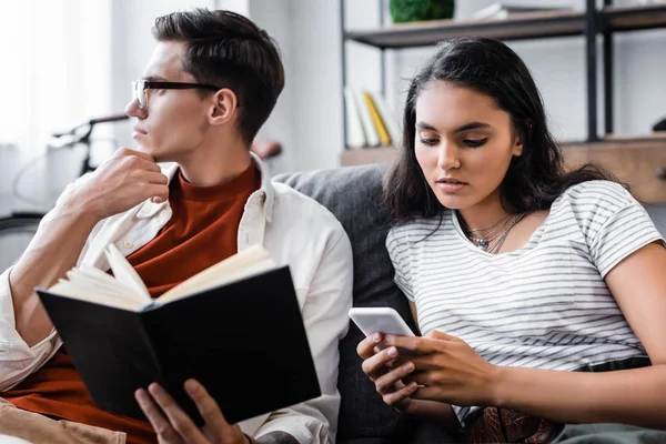 Étudiants multiculturels tenant un livre et utilisant un smartphone dans l'appartement — Photo de stock