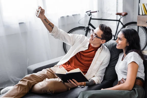 Étudiants multiculturels tenant un livre et prenant selfie dans l'appartement — Photo de stock