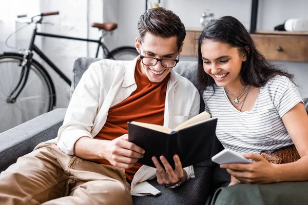 Feliz, estudantes multiculturais sorrindo e lendo livro no apartamento — Fotografia de Stock