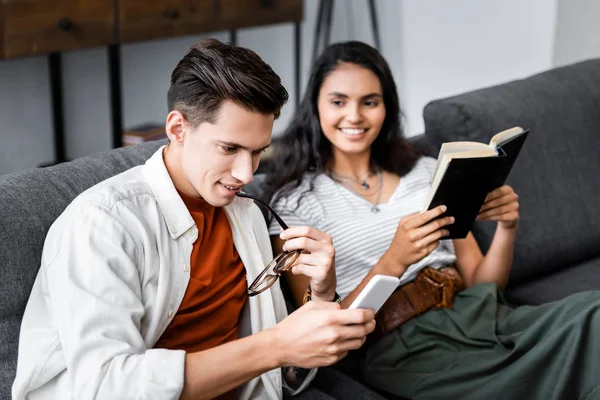Étudiants multiculturels tenant un livre et utilisant un smartphone dans l'appartement — Photo de stock