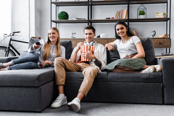 Amigos multiculturales sentados en el sofá y comiendo palomitas de maíz en el apartamento - foto de stock