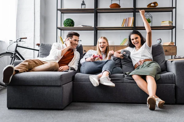 KYIV, UKRAINE - JULY 10, 2019: multicultural friends sitting on sofa and playing video game in apartment — Stock Photo